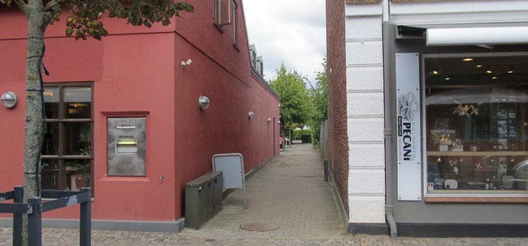 Åben passage langs med Andelskassen (den røde bygning) på billedet set fra Nørregade. Passagen foreslås styrket visuelt med pergola af virer med slyngplanter, belysning og belægning af røde klinker.